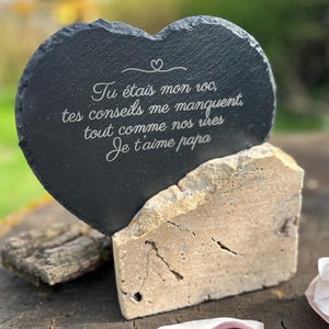 May include: A heart-shaped black slate plaque with rough, natural edges, engraved with the French text: Tu étais mon roc, tes conseils me manquent, tout comme nos rires, Je t'aime papa. The plaque is supported by a rectangular beige stone base.