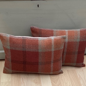 Puede incluir: Dos cojines rectangulares con un estampado de cuadros en tonos naranja, rojo y beige. Los cojines están colocados sobre un suelo de madera clara frente a un arcón de madera gris claro.