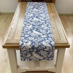 Può includere: Un runner da tavolo blu e bianco con un design floreale e di uccelli. Il runner è su un tavolo di legno.