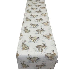 Può includere: Una tovaglia bianca con un motivo ripetuto di lepri ad acquerello in varie pose. Le lepri sono marroni e grigie, su uno sfondo di sottili pennellate blu e grigie. La tovaglia è realizzata in tessuto morbido.