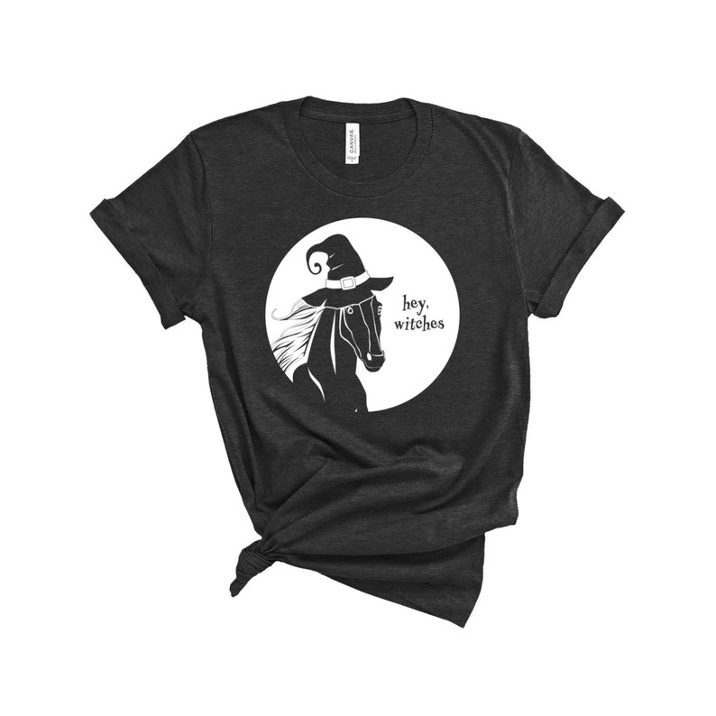 May include: A dark grey t-shirt with a white circle graphic featuring a black horse wearing a witch's hat. The text "hey, witches" is inside the circle.