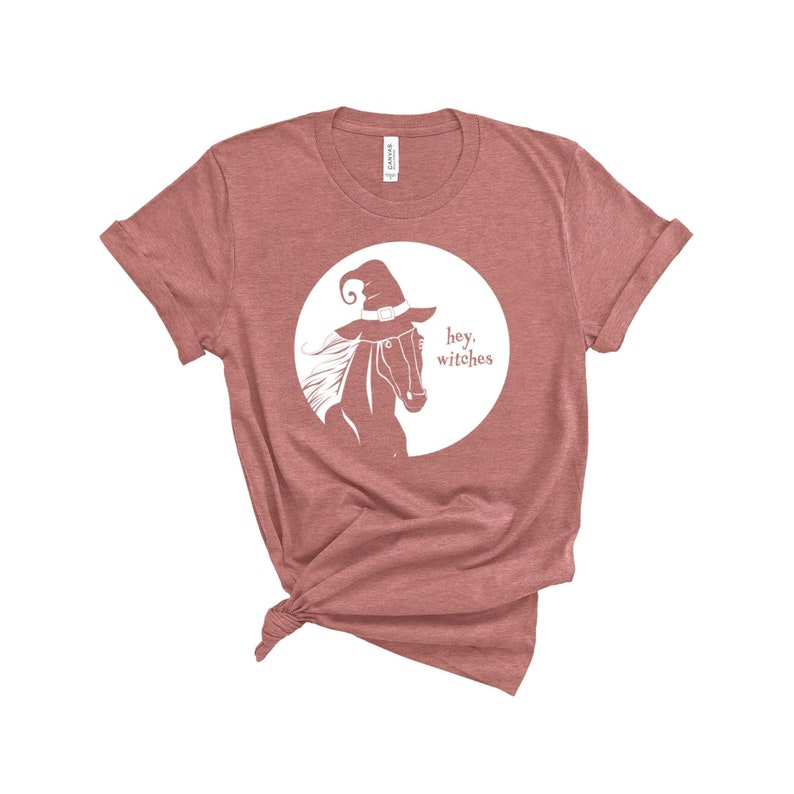 May include: A mauve colored t-shirt with a white graphic of a horse wearing a witch's hat and the text "hey, witches".