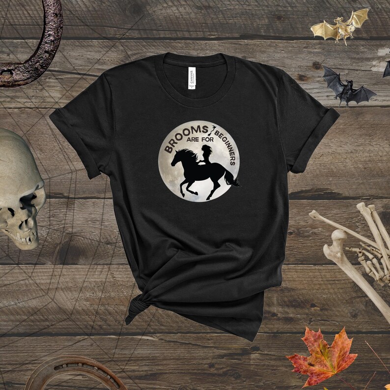 May include: Black t-shirt with a graphic of a witch riding a horse in front of a full moon. The text "Brooms are for beginners" is written around the moon.