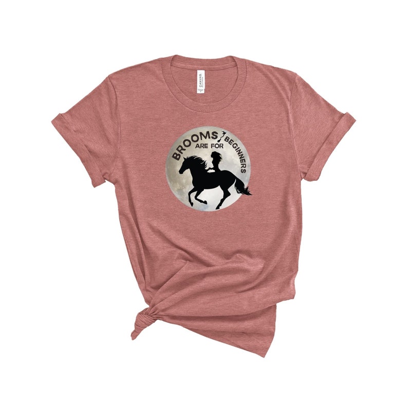 May include: A heather pink t-shirt with a graphic of a silhouette of a woman riding a horse on a moon with the text "Brooms are for beginners".