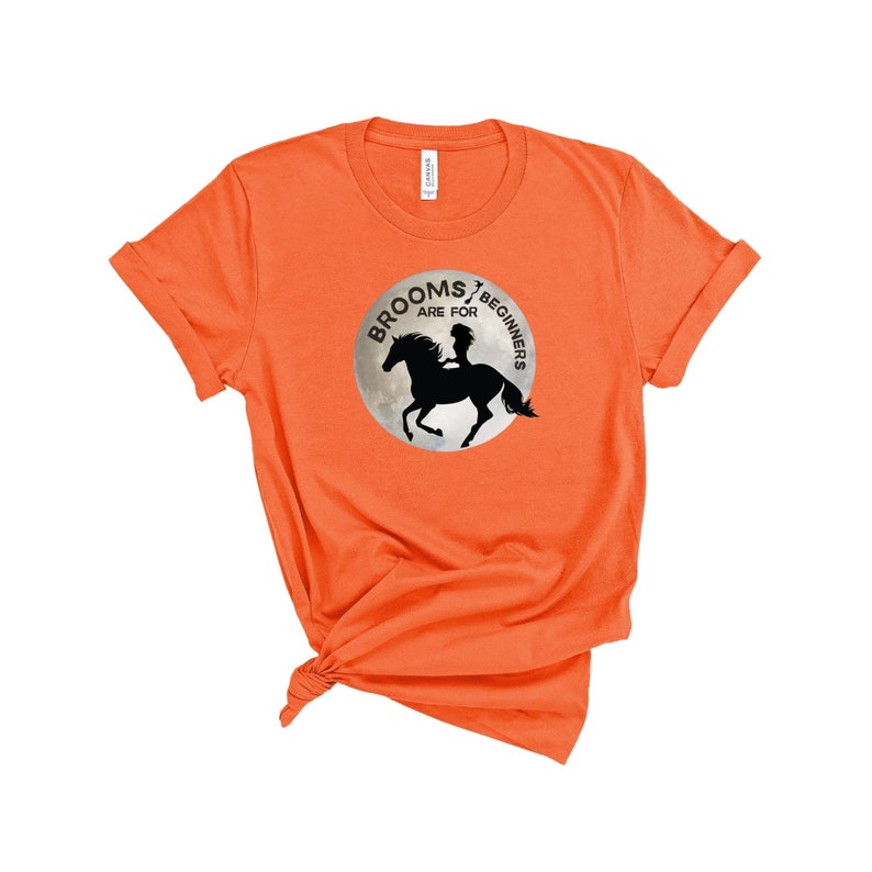 May include: Orange t-shirt with a black and white graphic of a witch riding a horse on a full moon. The graphic says "Brooms are for beginners".