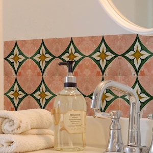 May include: A bathroom sink with a chrome tap, a clear soap dispenser, and a stack of white towels. The wall features pink tiles with green and yellow geometric patterns. A mirror is visible in the background.