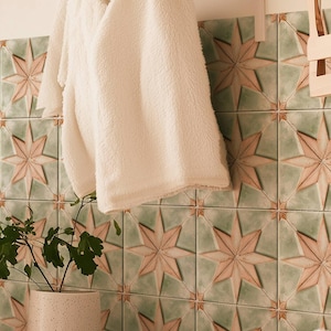 Puede incluir: Una toalla blanca y esponjosa colgada de un toallero en un baño con azulejos en forma de estrella verde y rosa. Hay toallas blancas apiladas en un estante debajo del toallero.