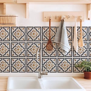 May include: Kitchen backsplash with decorative tiles featuring a floral design in gold and black. A white double sink and wooden countertops are in the foreground. Wooden shelves hold plants and kitchen utensils.