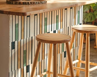 May include: A kitchen island with a wooden work surface and a decorative front with vertical stripes in green, beige, and black. Two wooden stools with round seats and angled legs stand in front. Potted plants are on the countertop.