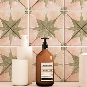 May include: Three bottles of bathroom products on a white countertop with a light wood shelf. The bottles are a white pump bottle, a brown glass bottle with a pump, and a white ceramic cup. The bottles are in front of a wall with pink and green star-shaped tiles.