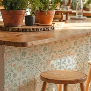 Puede incluir: Una isla de cocina con azulejos cuadrados que presentan un patrón floral repetitivo en tonos verde y melocotón apagados. La isla tiene una encimera de madera gruesa con dos macetas de terracota con plantas verdes y una pequeña maceta de cerámica azul.