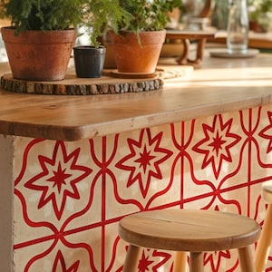 May include: A kitchen island with a wooden countertop and a decorative front featuring a repeating red star pattern on a cream background. Terra cotta pots with green plants sit on the countertop, along with a small wooden stool.
