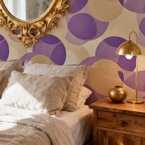 Peut inclure: Papier peint avec un motif répétitif de cercles superposés dans des tons de violet et de beige. Le design recouvre le mur derrière un lit avec une literie claire, une table de chevet en bois, une lampe en laiton et un miroir doré orné.