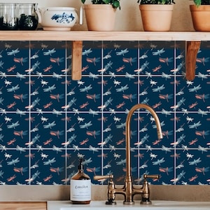 May include: A set of square wall tiles featuring a repeating pattern of dragonflies in shades of white, light blue, and coral pink against a dark navy blue background, installed as a backsplash behind a kitchen sink and faucet.