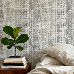 Op de afbeelding: Een slaapkamer met een kleine potplant op een stapel boeken op een houten nachtkastje. Het bed heeft beige lakens en kussens. Een zwarte koptelefoon ligt op het bed. De muur heeft een textuur, geweven patroon.