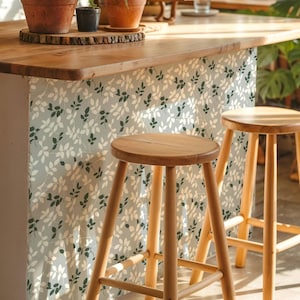 May include: A kitchen island with a light wood worktop and a leaf-patterned facade. Two wooden stools with round seats and angled legs stand in front of the island. Potted plants sit on the worktop, adding a touch of greenery.