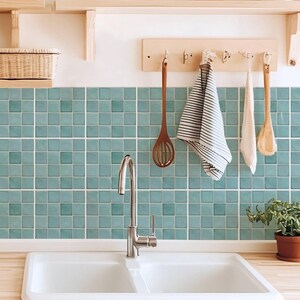May include: A bright kitchen scene featuring a white double sink with a chrome faucet. Teal square tiles form a backsplash. Wooden shelves hold plants and a basket. Wooden utensils and a striped towel hang from a wooden rack.
