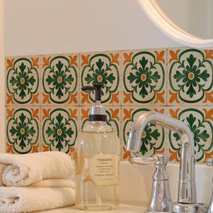 May include: Bathroom sink with a chrome faucet, a clear soap dispenser, and a stack of white towels. Decorative tiles with green and orange floral patterns adorn the wall. A round mirror is visible in the background.