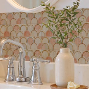 May include: A bathroom vanity area featuring a wall with square tiles in a repeating fan-shaped pattern of soft pink, cream, and gold. A chrome tap, a cream-coloured ceramic vase with green leafy branches, and a wooden tray with a lemon wedge.