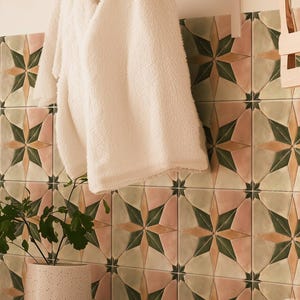 May include: A fluffy white towel hangs on a rack against a backdrop of patterned tiles. The tiles feature a starburst design in shades of green, pink, and beige. A potted plant and folded towels are also visible.