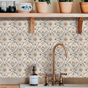 Pode incluir: Interior da cozinha com uma pia branca, torneira de latão e backsplash de azulejos estampados. O azulejo apresenta um fundo creme com desenhos ornamentais dourados e bege. Um dispensador de sabonete e plantas em vasos também são visíveis.