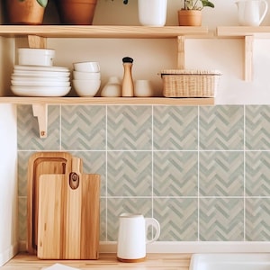 May include: A kitchen interior with wooden shelves displaying potted plants, white crockery, and a wicker basket. Below, a wooden worktop holds chopping boards, a white jug, and a sink. The backsplash has a light blue and grey chevron pattern.