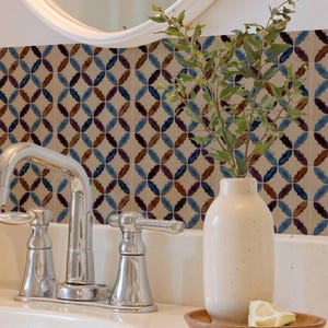 May include: A cream-colored ceramic vase sits on a small wooden tray next to a chrome bathroom faucet. The vase contains a green leafy branch, and a slice of lemon rests on the tray. The background features a wall with a repeating tile pattern.