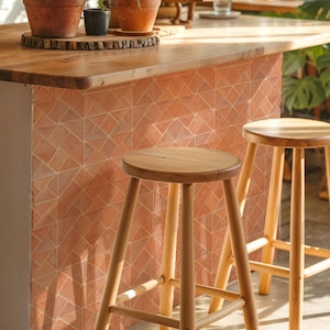 Op de afbeelding: Een keukeneiland met een houten aanrechtblad en een terracotta tegelgevel. Twee houten krukken staan voor het eiland. Potplanten staan op het aanrechtblad en geven de ruimte groen.