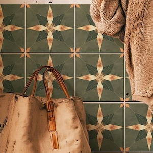 Peut inclure: Un sac fourre-tout en toile avec des poignées en cuir est posé contre un mur de carreaux verts. Les carreaux présentent un motif d'éclat d'étoile en or et crème. Un pull beige est accroché à proximité, et une couverture blanche pliée est en bas.