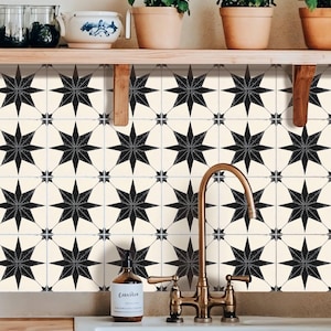 May include: A kitchen backsplash featuring a repeating pattern of black starbursts on a cream backdrop. The design is a modern take on a classic tile pattern. A brass tap and white sink are in the foreground, with plants on a shelf above.