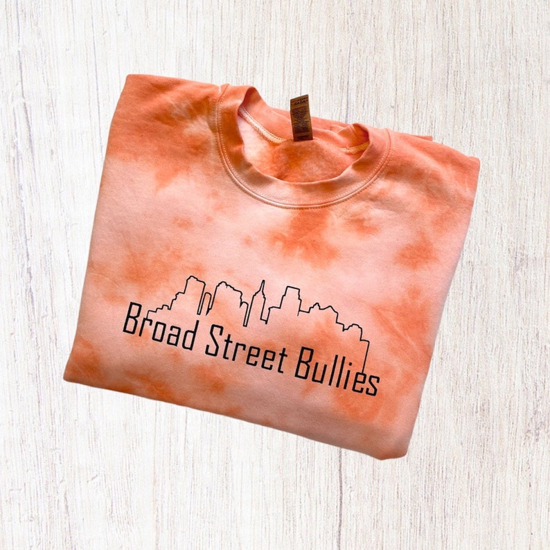 Pode incluir: Camisola laranja tie-dye com um contorno preto de um horizonte da cidade e o texto "Broad Street Bullies".