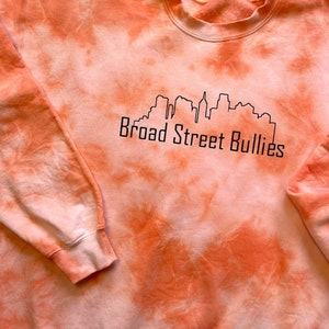 Pode incluir: Camisola laranja tie-dye com um contorno preto de um horizonte da cidade e o texto "Broad Street Bullies".