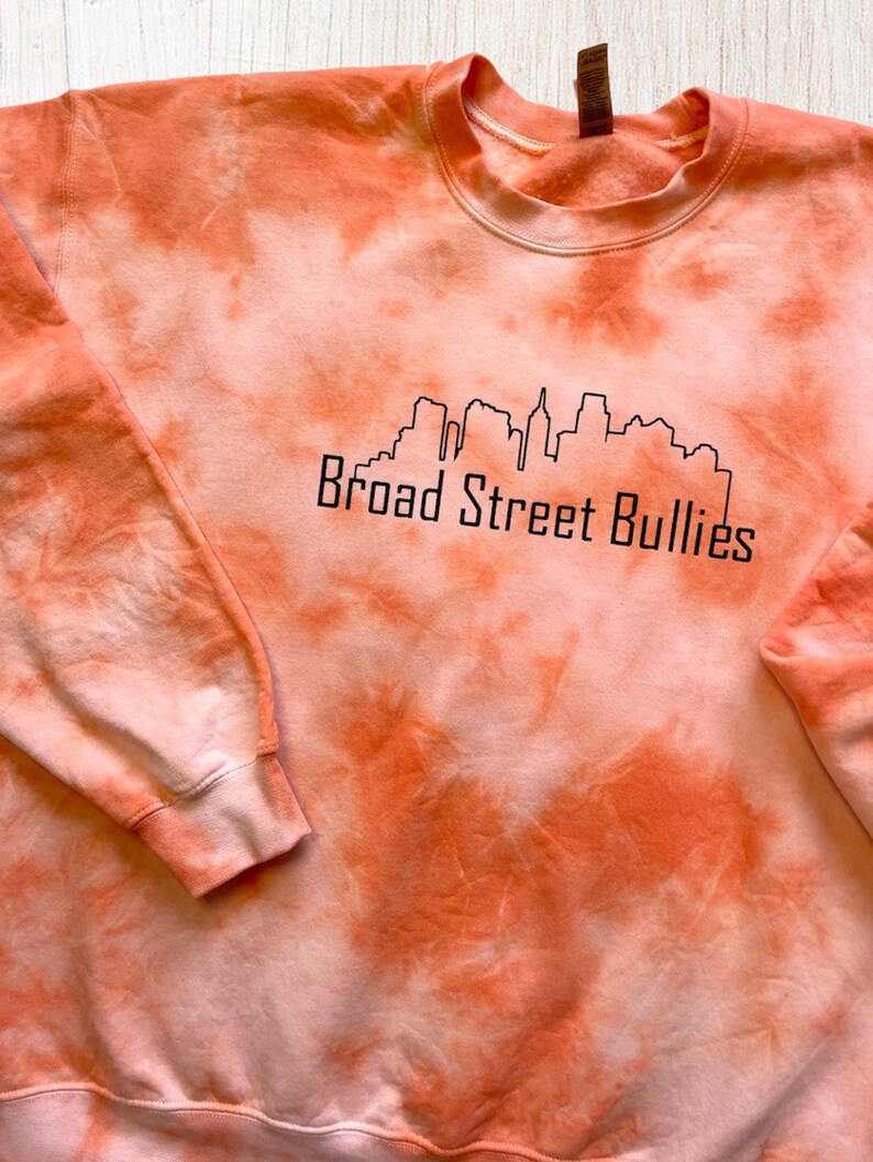 Pode incluir: Camisola laranja tie-dye com um contorno preto de um horizonte da cidade e o texto "Broad Street Bullies".
