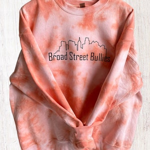 Pode incluir: Camisola laranja tie-dye com um contorno preto de um horizonte da cidade e o texto "Broad Street Bullies".