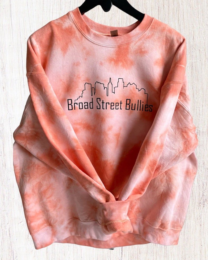 Pode incluir: Camisola laranja tie-dye com um contorno preto de um horizonte da cidade e o texto "Broad Street Bullies".