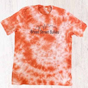 Pode incluir: Camiseta laranja e branca tie-dye com um contorno preto de um horizonte da cidade e o texto "Broad Street Bullies".