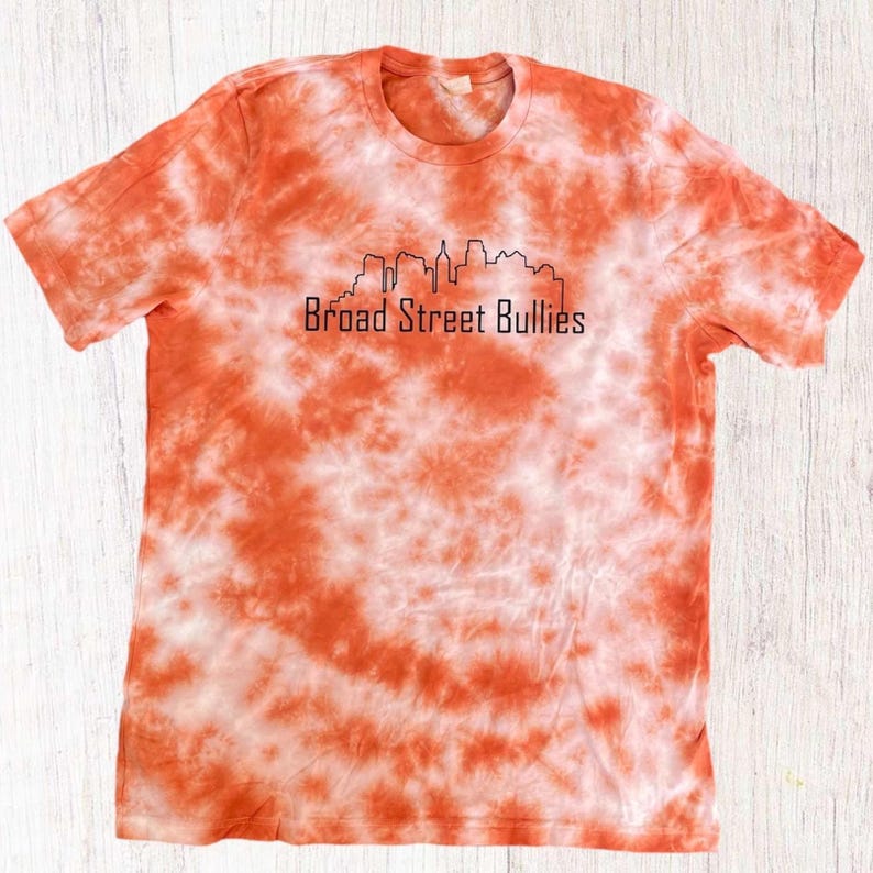 Pode incluir: Camiseta laranja e branca tie-dye com um contorno preto de um horizonte da cidade e o texto "Broad Street Bullies".
