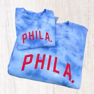 May include: Two blue tie-dye sweatshirts with the word "PHILA." printed in red on the front. The sweatshirts are folded on top of each other.