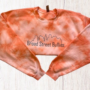 Pode incluir: Camisola laranja tie-dye com um contorno preto de um horizonte da cidade e o texto "Broad Street Bullies".