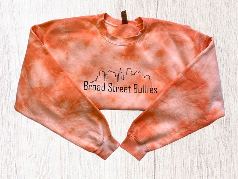 Pode incluir: Camisola laranja tie-dye com um contorno preto de um horizonte da cidade e o texto "Broad Street Bullies".