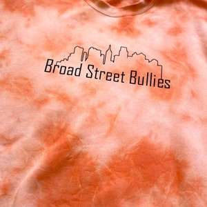 Pode incluir: Camisola laranja tie-dye com um contorno preto de um horizonte da cidade e o texto "Broad Street Bullies".
