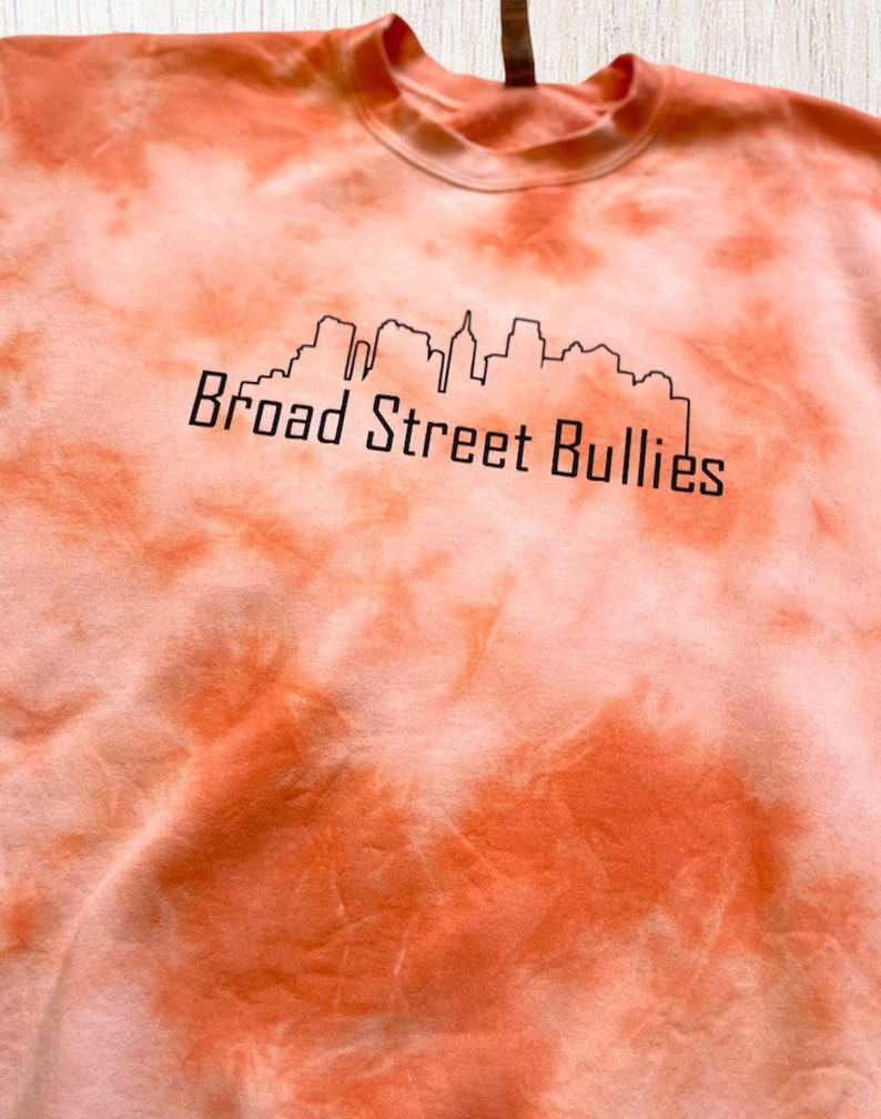 Pode incluir: Camisola laranja tie-dye com um contorno preto de um horizonte da cidade e o texto "Broad Street Bullies".