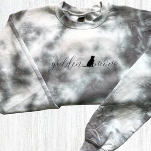 Pu&ograve; includere: Una felpa grigia tie-dye con il testo "golden mom" e la silhouette di un golden retriever.