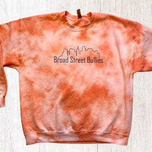 Pode incluir: Camisola laranja tie-dye com um contorno preto de um horizonte da cidade e o texto "Broad Street Bullies".