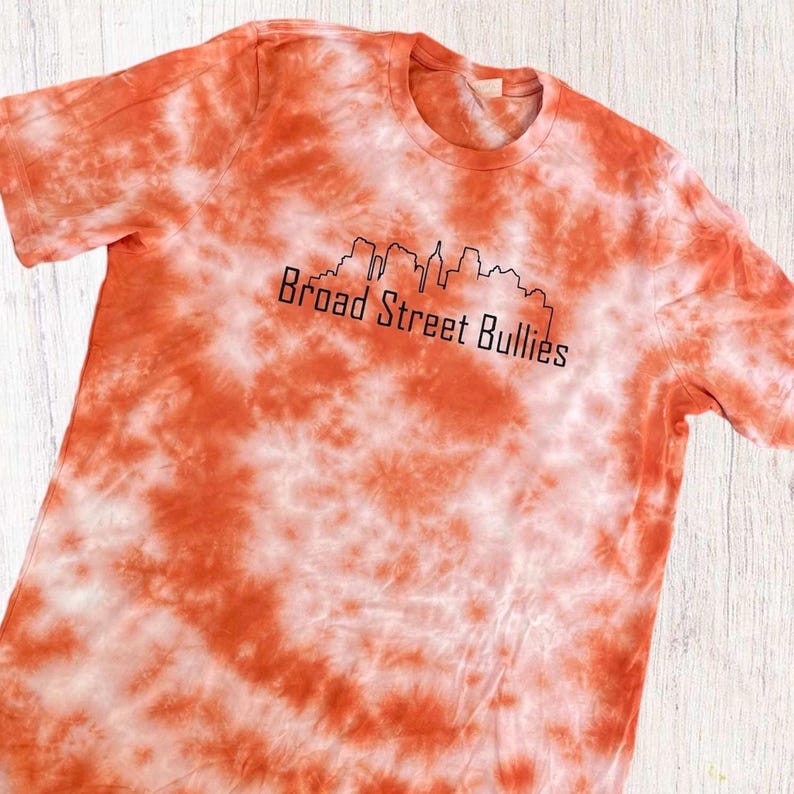 Pode incluir: Camiseta laranja e branca tie-dye com um contorno preto de um horizonte da cidade e o texto "Broad Street Bullies".