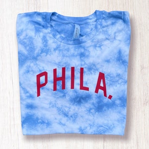 May include: A blue tie-dye t-shirt with the word "PHILA." printed in red on the front.