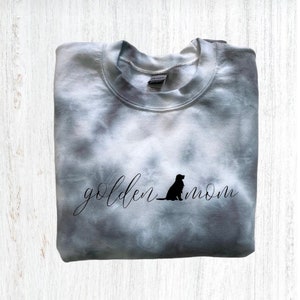 Pu&ograve; includere: Una felpa grigia tie-dye con il testo "golden mom" in scrittura nera e una silhouette nera di un golden retriever seduto.