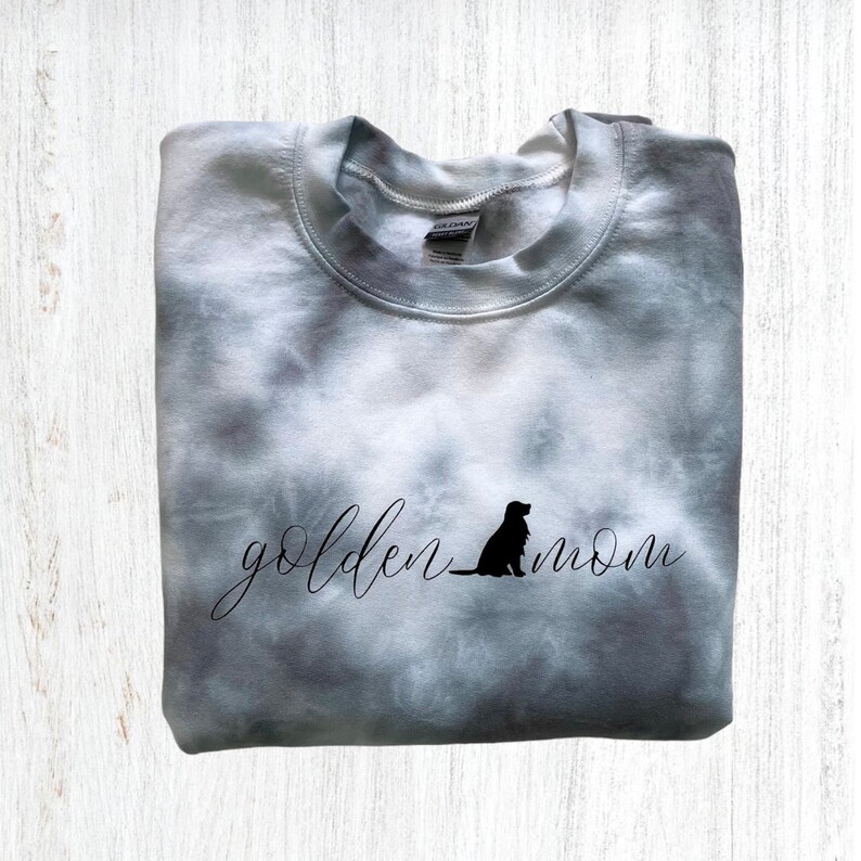 Pu&ograve; includere: Una felpa grigia tie-dye con il testo "golden mom" in scrittura nera e una silhouette nera di un golden retriever seduto.