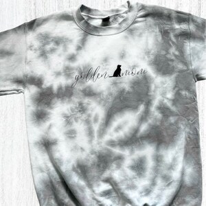 Pu&ograve; includere: Una felpa grigia tie-dye con il testo "golden mom" e la silhouette di un golden retriever.