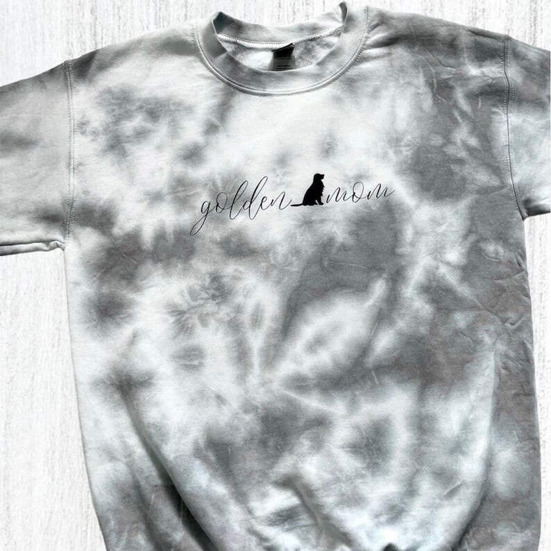 Pu&ograve; includere: Una felpa grigia tie-dye con il testo "golden mom" e la silhouette di un golden retriever.